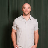 Man wearing a light gray polo shirt against a dark green curtain
