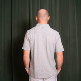 Man wearing a gray patterned shirt and light-colored pants against a dark green curtain.