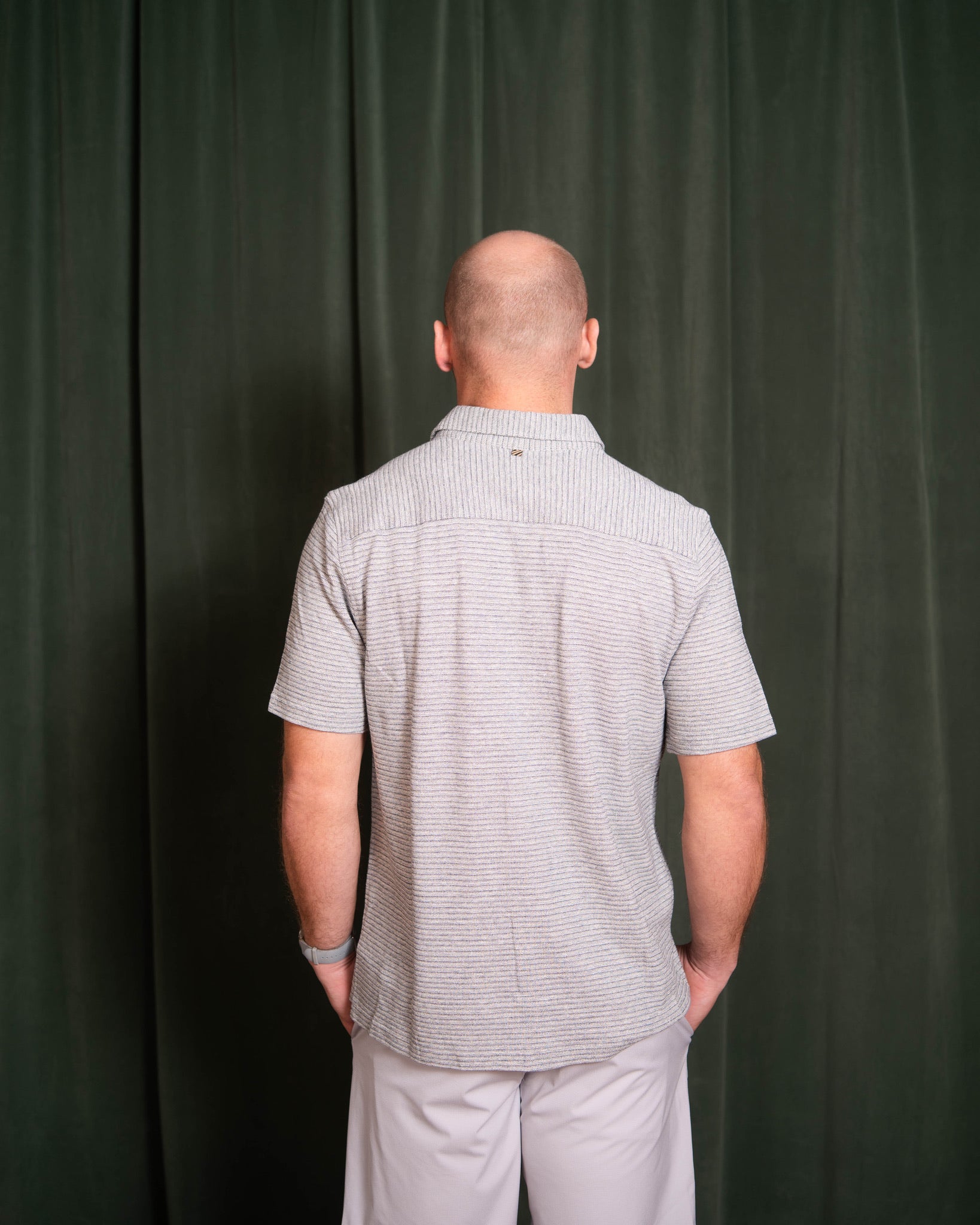 Man wearing a gray patterned shirt and light-colored pants against a dark green curtain.