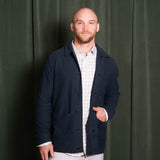 Man wearing a navy jacket over a white shirt against a green curtain background