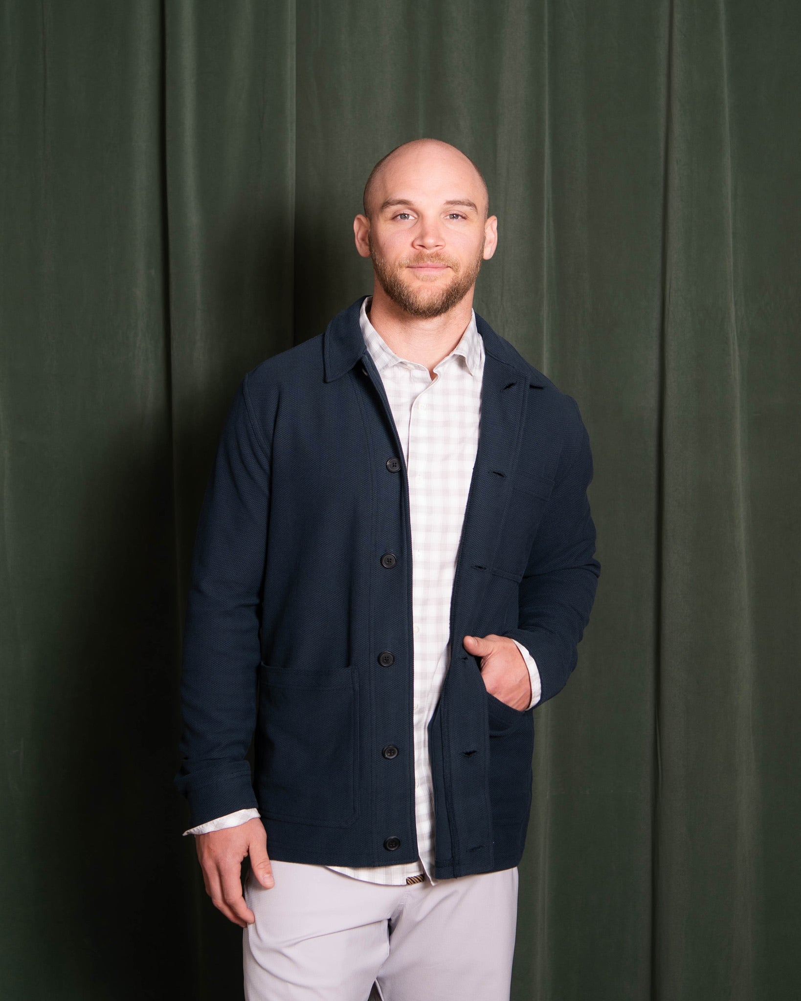 Man wearing a navy jacket over a white shirt against a green curtain background