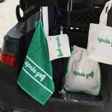 A set of golf-themed reusable bags and towels hanging from a golf bag in a cart. The items include a green towel with a white stripe and script reading 'après golf', a small white pouch, a larger white drawstring bag with green trim and script, and a white tote bag with green script.